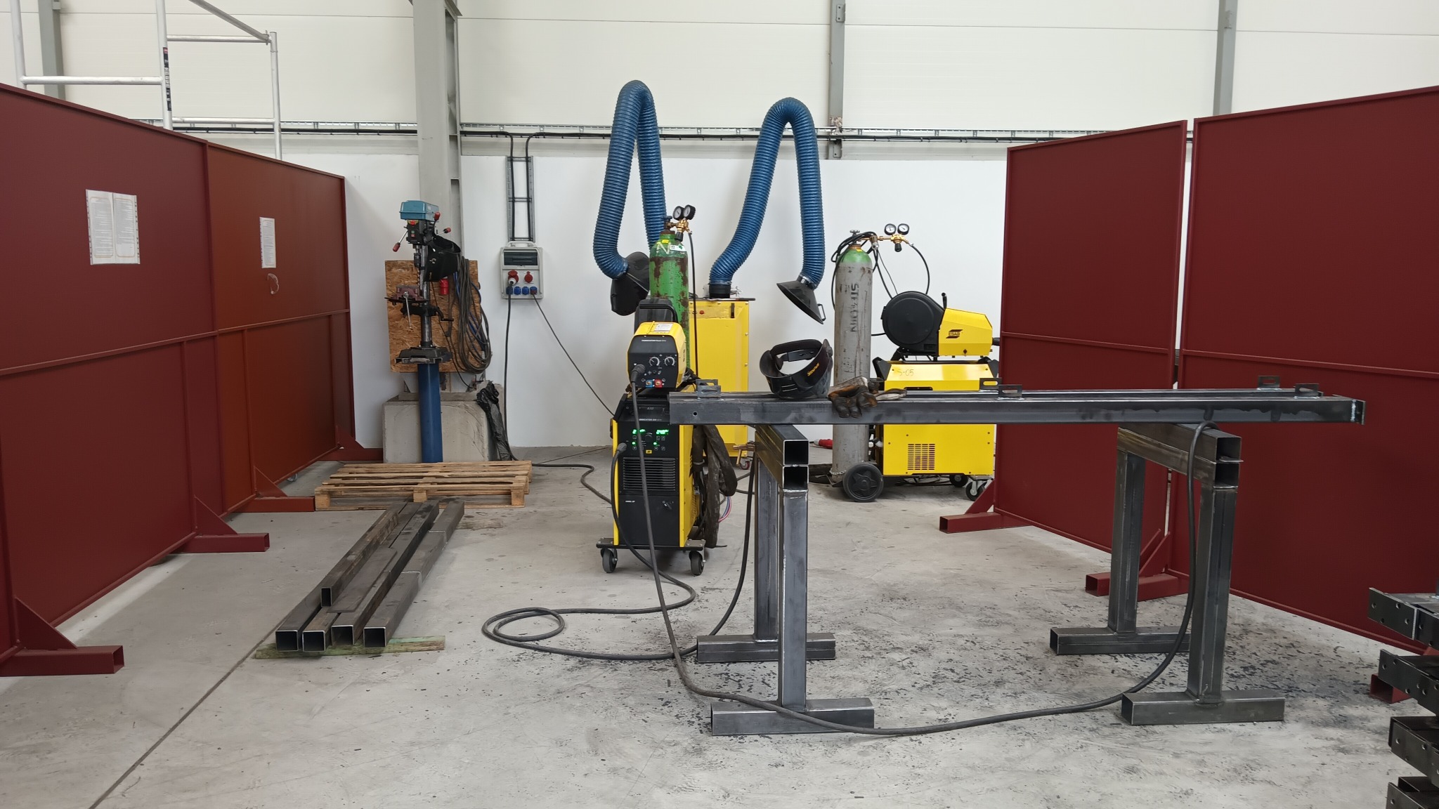 Warsztat spawalniczy z żółtymi spawarkami, metalowymi konstrukcjami, osłonami spawalniczymi i systemem odciągu dymu. Widoczne elementy metalowe w trakcie obróbki.