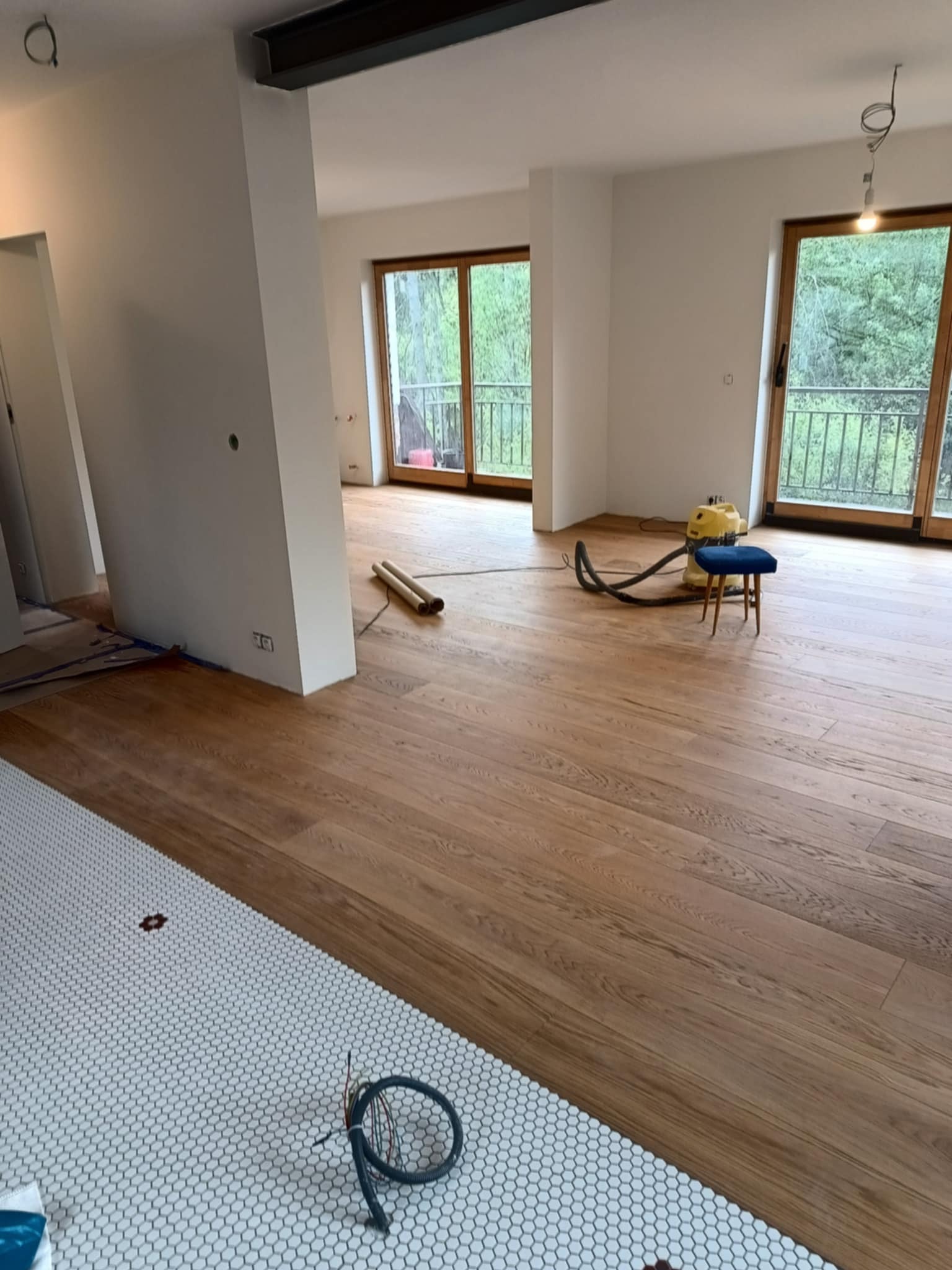 Jasne wnętrze w trakcie prac wykończeniowych: parkiet po renowacji, fragment mozaiki podłogowej i odkurzacz przemysłowy. Widok na okna z zielenią w tle.