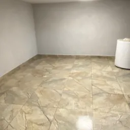 Beżowe płytki podłogowe imitujące marmur w pustym pomieszczeniu z szarymi ścianami i białym zbiornikiem na wodę w rogu. Widok na całą podłogę.