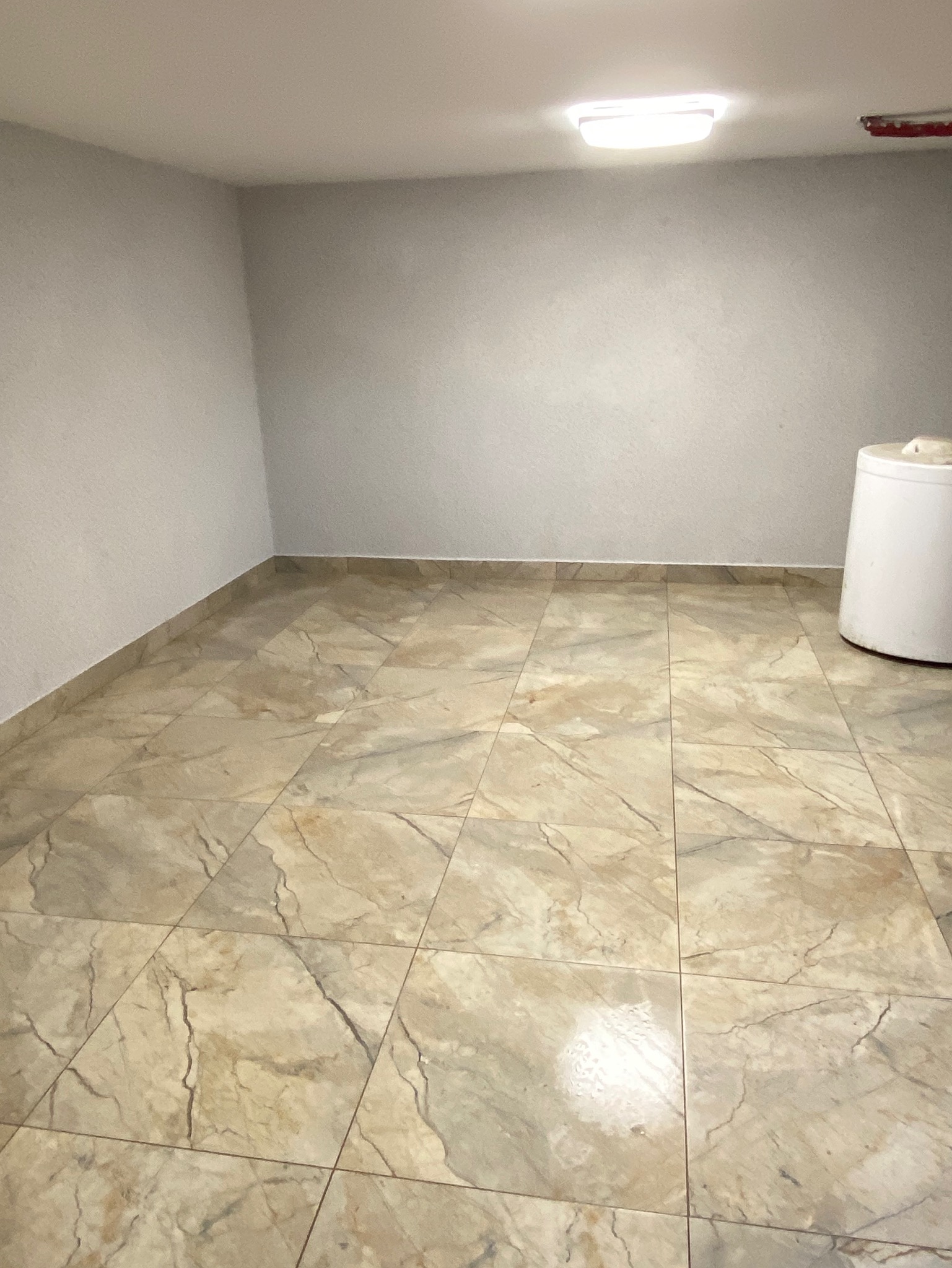 Beżowe płytki podłogowe imitujące marmur w pustym pomieszczeniu z szarymi ścianami i białym zbiornikiem na wodę w rogu. Widok na całą podłogę.