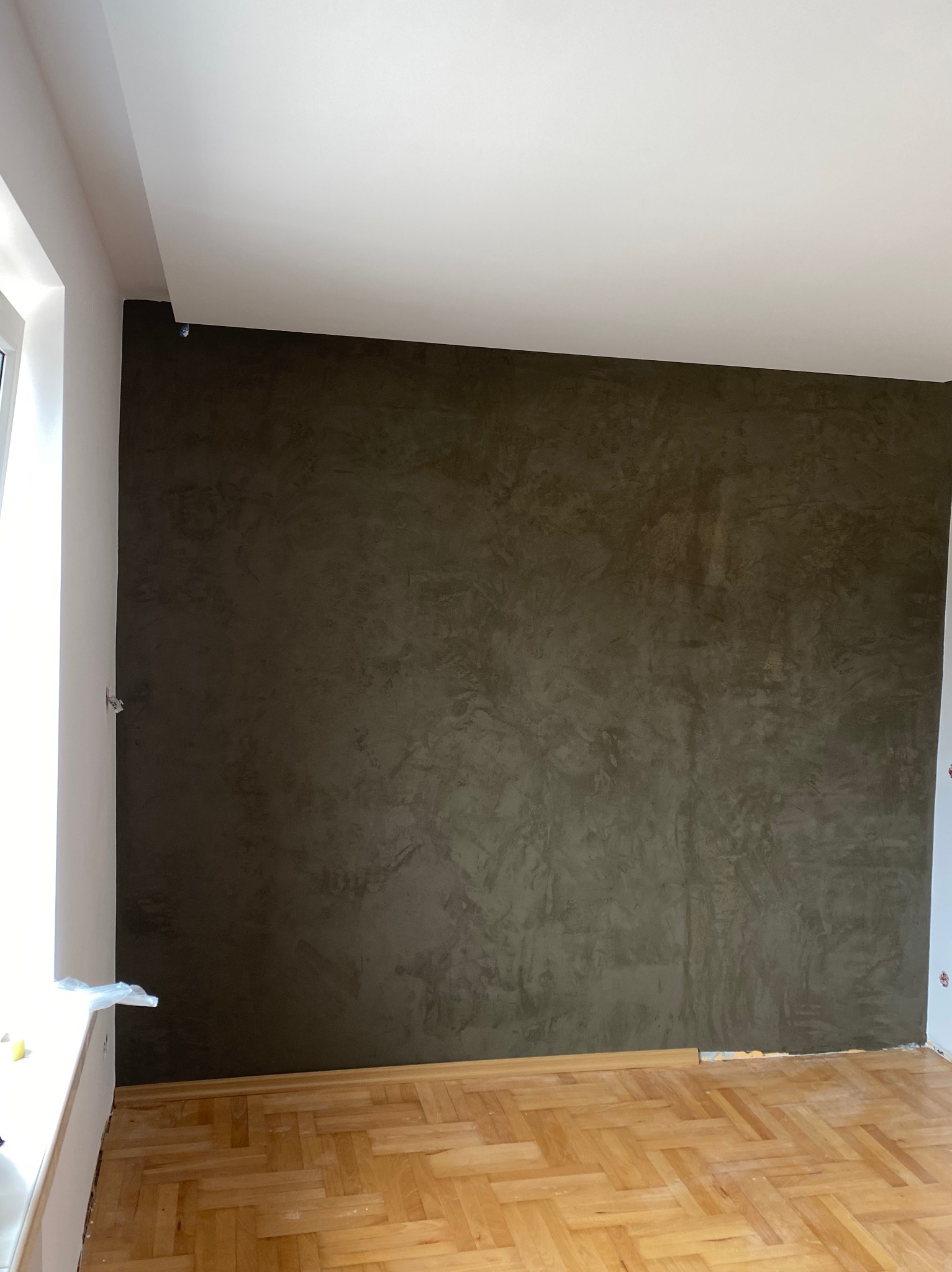 Ściana w odcieniach brązu z efektem dekoracyjnym, widoczna drewniana podłoga w jodełkę, fragment okna po lewej stronie.