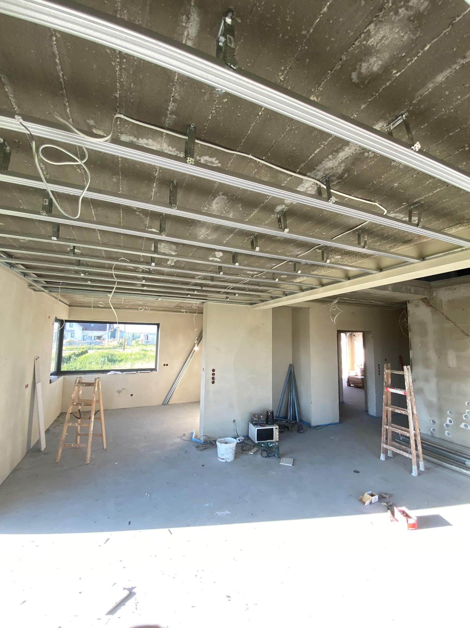 Wnętrze w trakcie montażu sufitu podwieszanego z widoczną konstrukcją metalową i instalacją elektryczną. W tle okno z widokiem na zieleń i drabina.