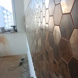 Ściana wykończona heksagonalnymi, metalicznymi płytkami. Widoczne narzędzia i okno w tle. Nietypowy wzór i materiał płytek. Ukończony fragment ściany.