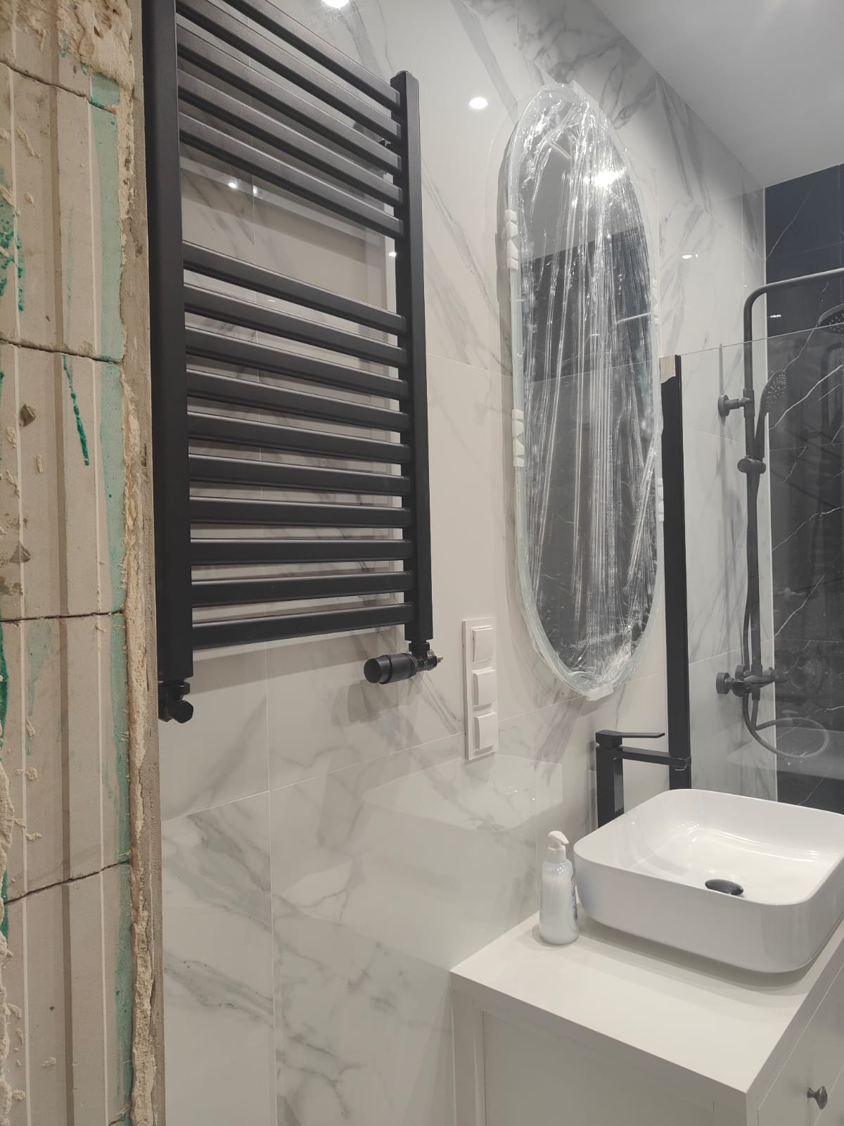 Częściowo wykończona łazienka z czarnym grzejnikiem, białą umywalką i owalnym lustrem w folii ochronnej na tle marmurowych płytek. Widoczny fragment ściany w trakcie prac.