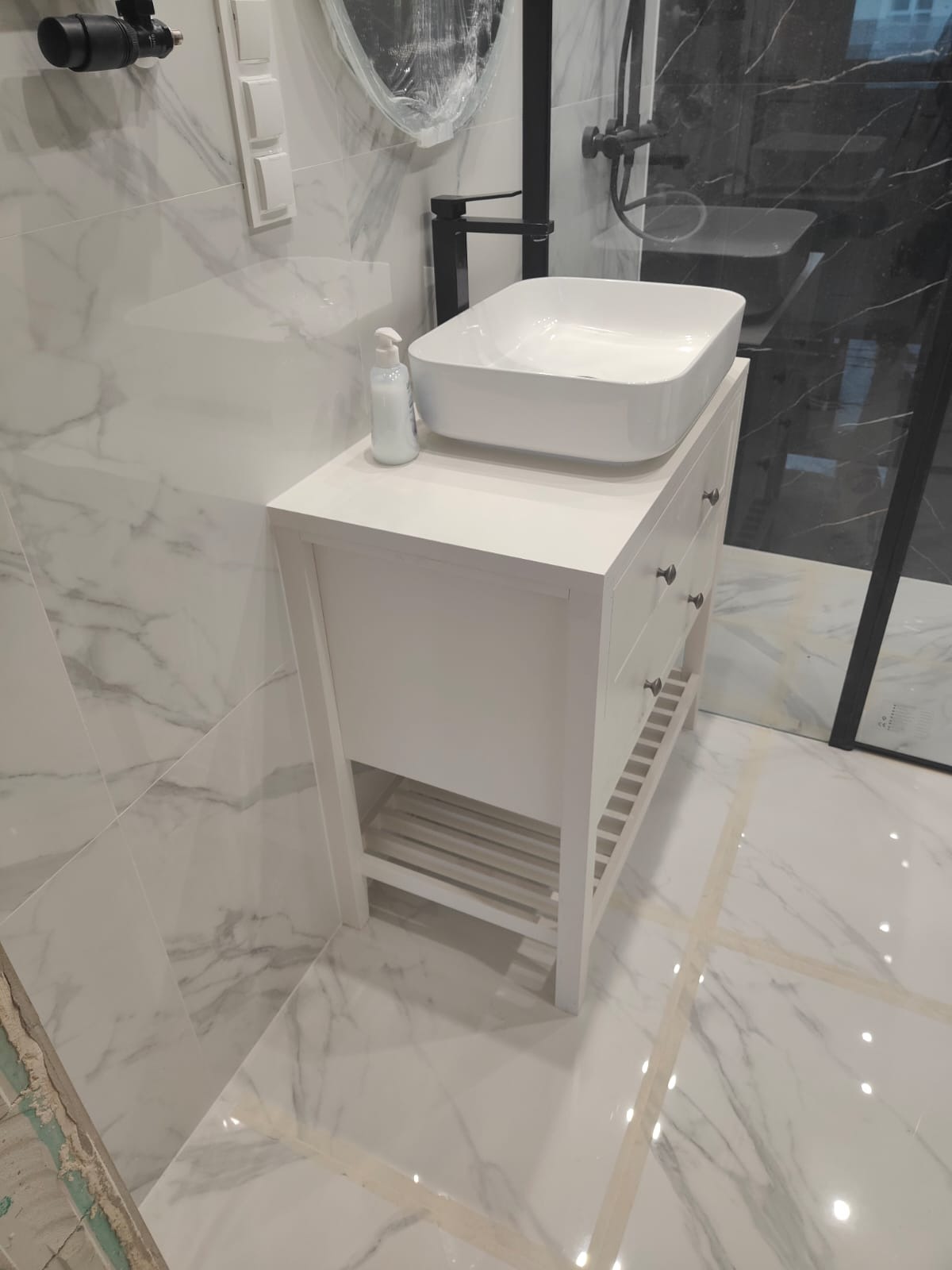 Biała szafka łazienkowa z umywalką nablatową, marmurowe płytki na ścianie i podłodze, czarna armatura, nowoczesny design, mydło w dozowniku.