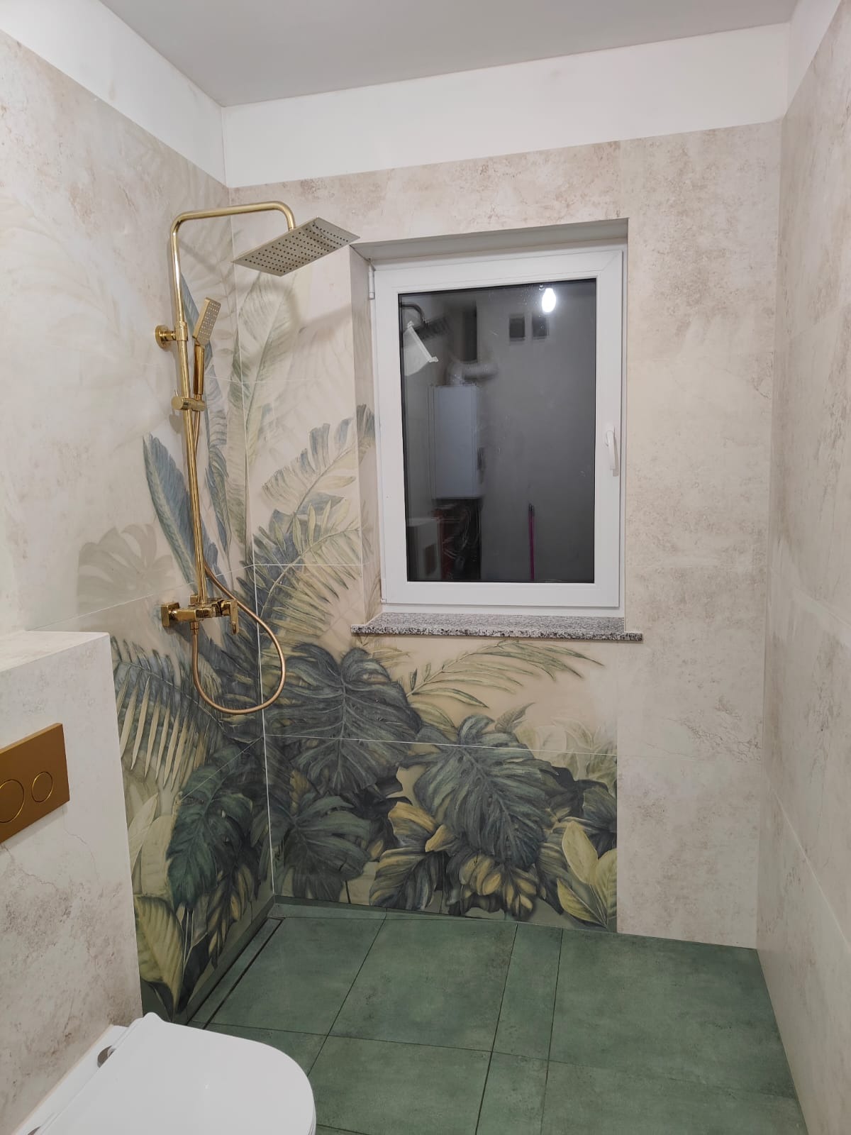 Wykończona łazienka z kabiną prysznicową w odcieniach beżu i zieleni. Na ścianach płytki z motywem roślinnym, złota armatura i okno z kamiennym parapetem.