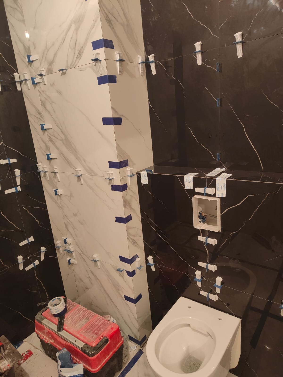 Biało-czarne płytki imitujące marmur, ułożone z użyciem klipsów i taśmy. W tle widoczna toaleta i czerwona skrzynka narzędziowa. Prace glazurnicze w toku.