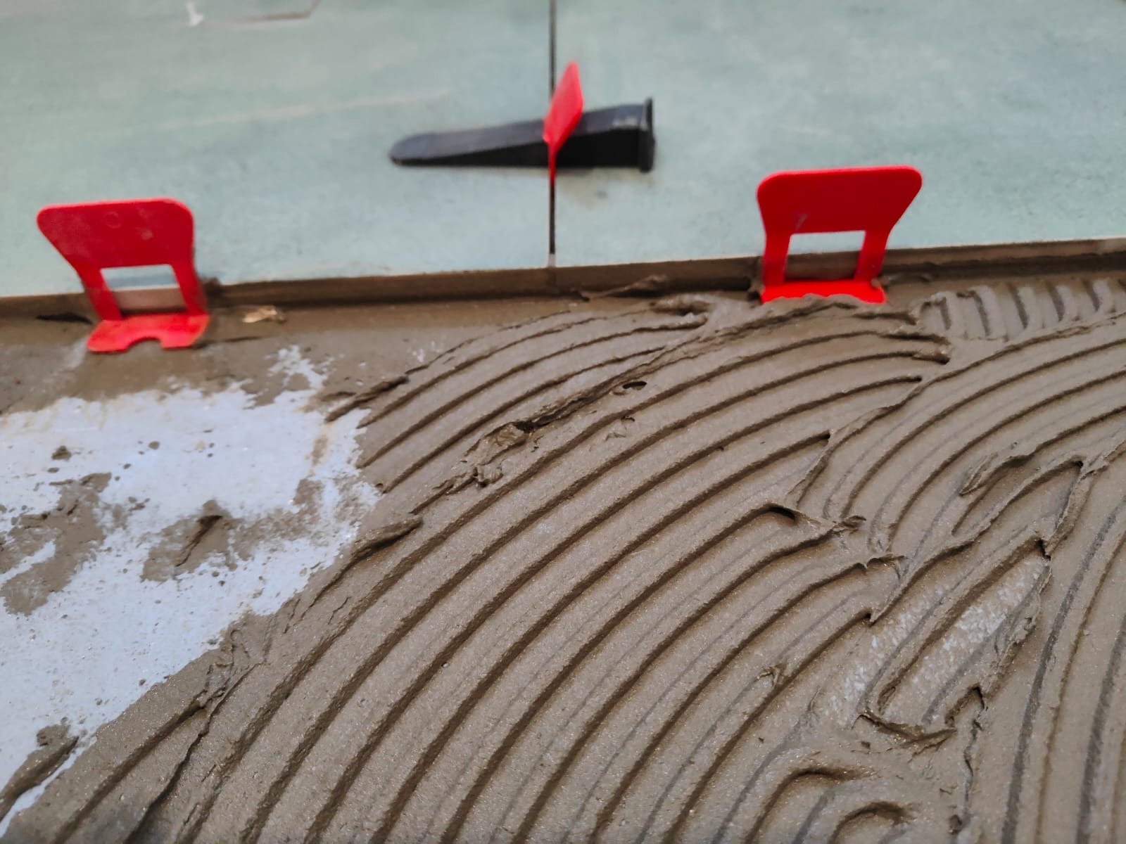 Świeżo nałożona warstwa kleju pod płytki, widoczne czerwone klipsy poziomujące i czarne kliny. Układanie płytek w trakcie realizacji z bliska.