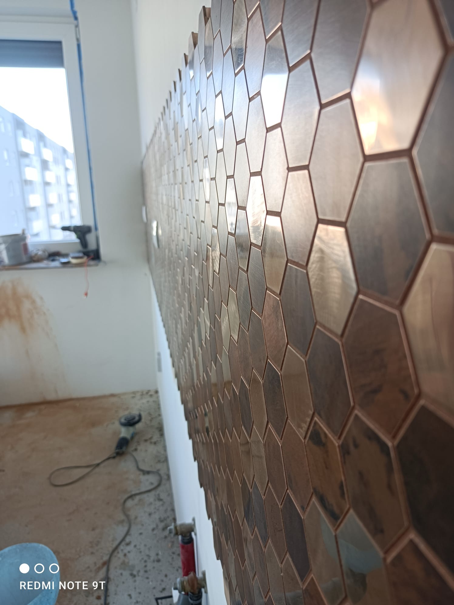 Ściana wykończona heksagonalnymi, metalicznymi płytkami. Widoczne narzędzia i okno w tle. Nietypowy wzór i materiał płytek. Ukończony fragment ściany.