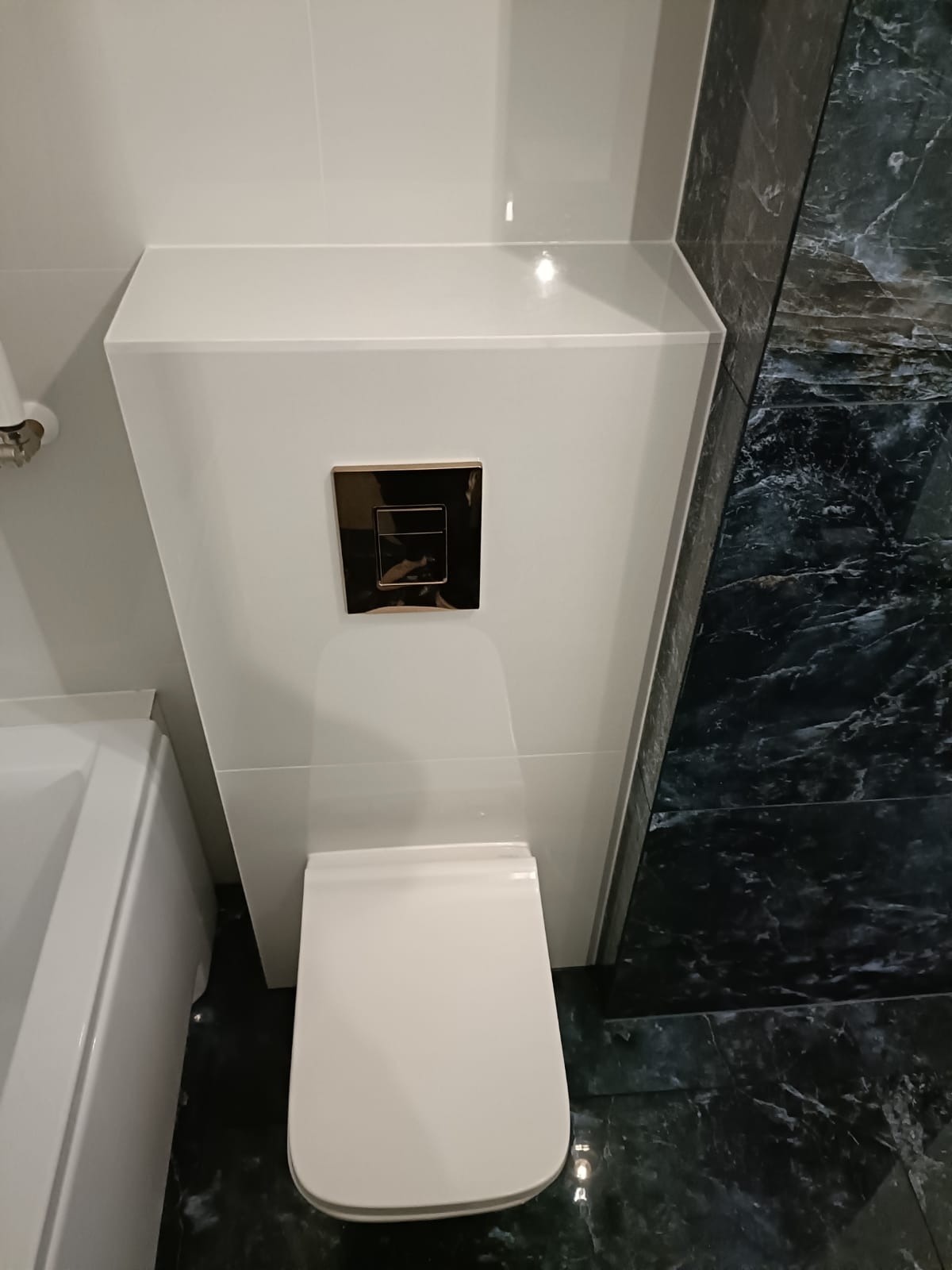 Nowoczesna łazienka: biała zabudowa toalety z chromowanym przyciskiem spłukiwania, marmurowe płytki podłogowe i ścienne. Minimalistyczny design, eleganckie wykończenie.