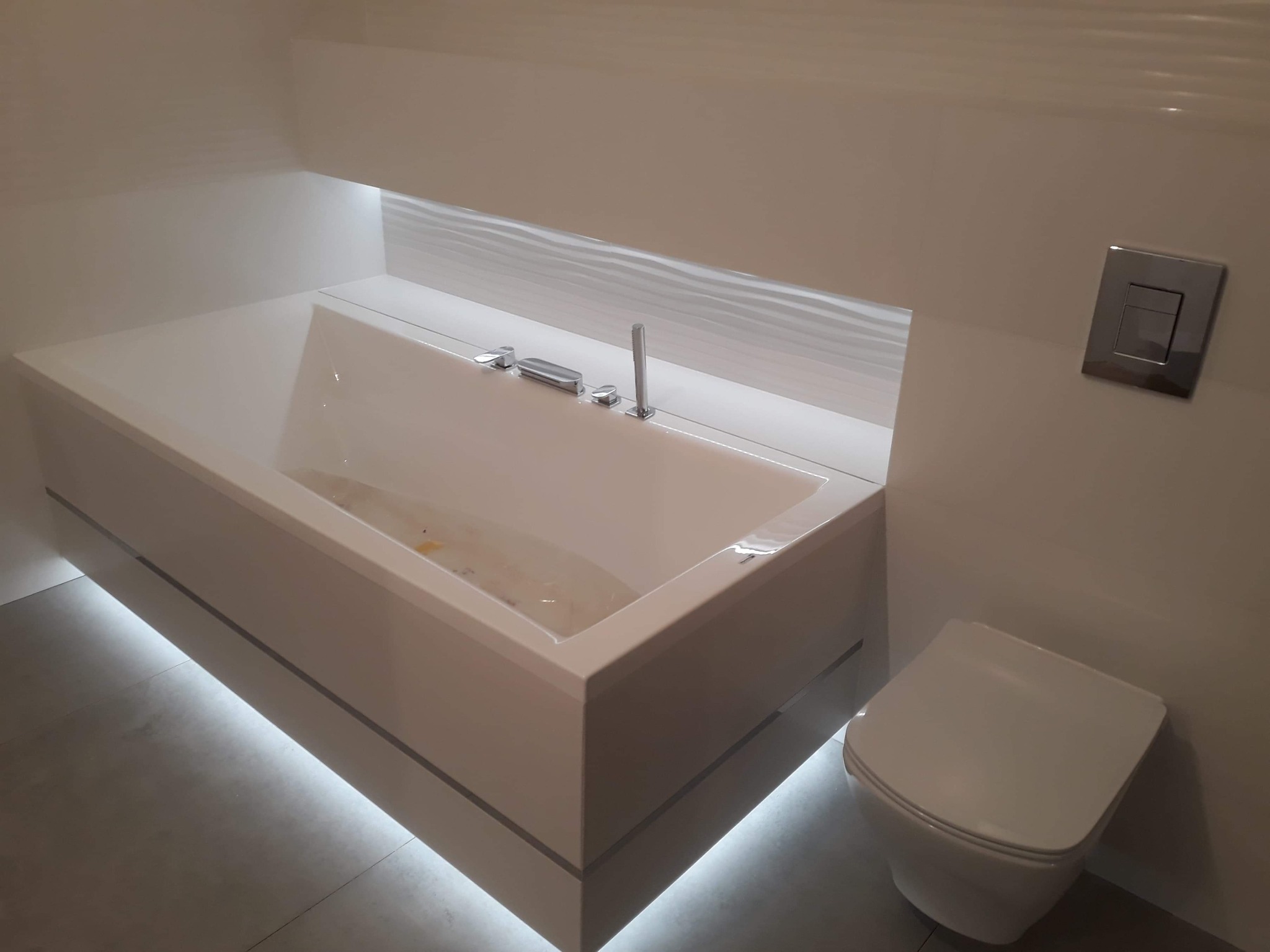 Nowoczesna łazienka z podświetlaną wanną, wiszącą toaletą i minimalistyczną armaturą. Ściany wykończone jasnymi płytkami, podkreślone subtelnym oświetleniem.