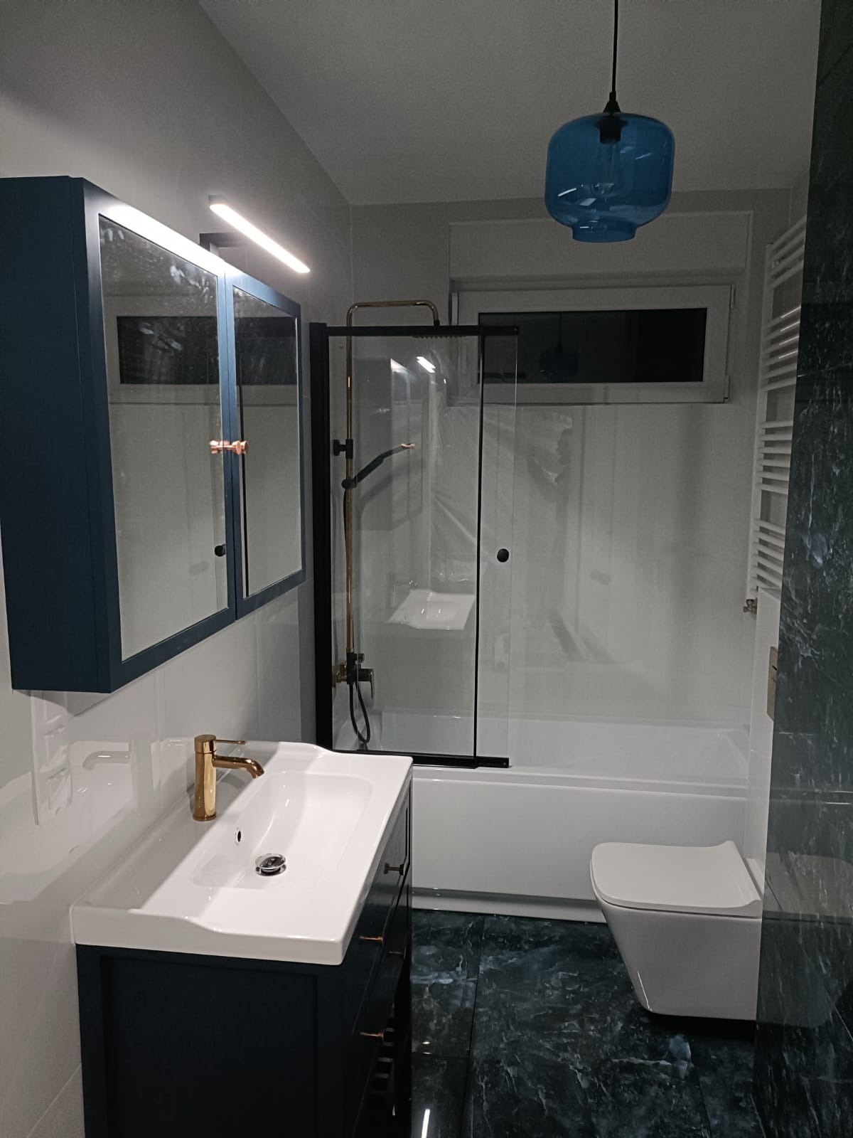Nowoczesna łazienka po remoncie: ciemne szafki z lustrami, złota armatura, kabina prysznicowa z czarnymi ramami i oryginalna, błękitna lampa wisząca.