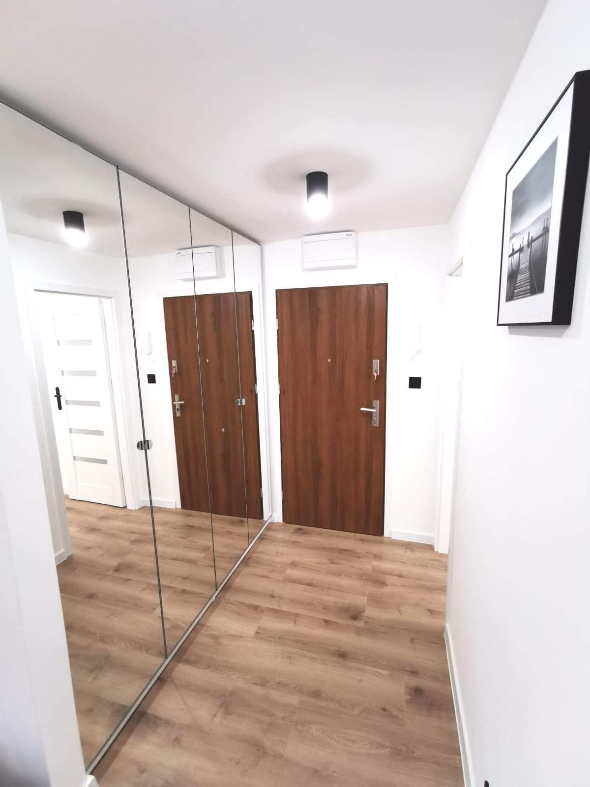 Wnętrze korytarza we Wrocławiu z drewnianą podłogą, lustrem na ścianie i dwojgiem drzwi. Minimalistyczny design z czarnymi akcentami i obrazem na ścianie.