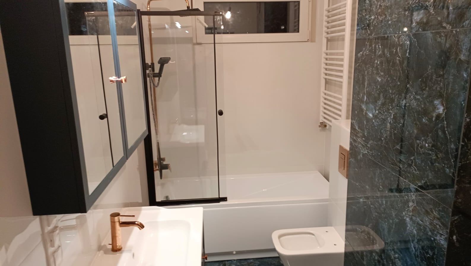 Nowoczesna łazienka z marmurową ścianą, kabiną prysznicową w czarnej ramie i armaturą w kolorze miedzi. Widoczny grzejnik drabinkowy i szafka z lustrem.