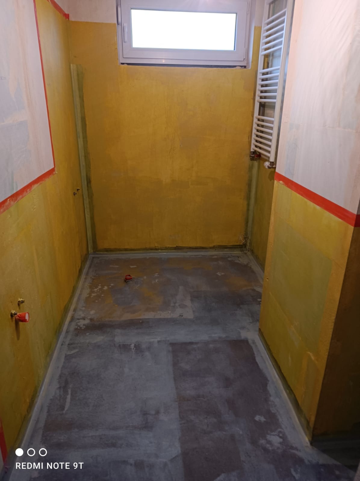 Przygotowana łazienka do dalszych prac: żółte ściany z taśmą, surowa podłoga, grzejnik drabinkowy i małe okno. Widoczne punkty instalacji wodnej.