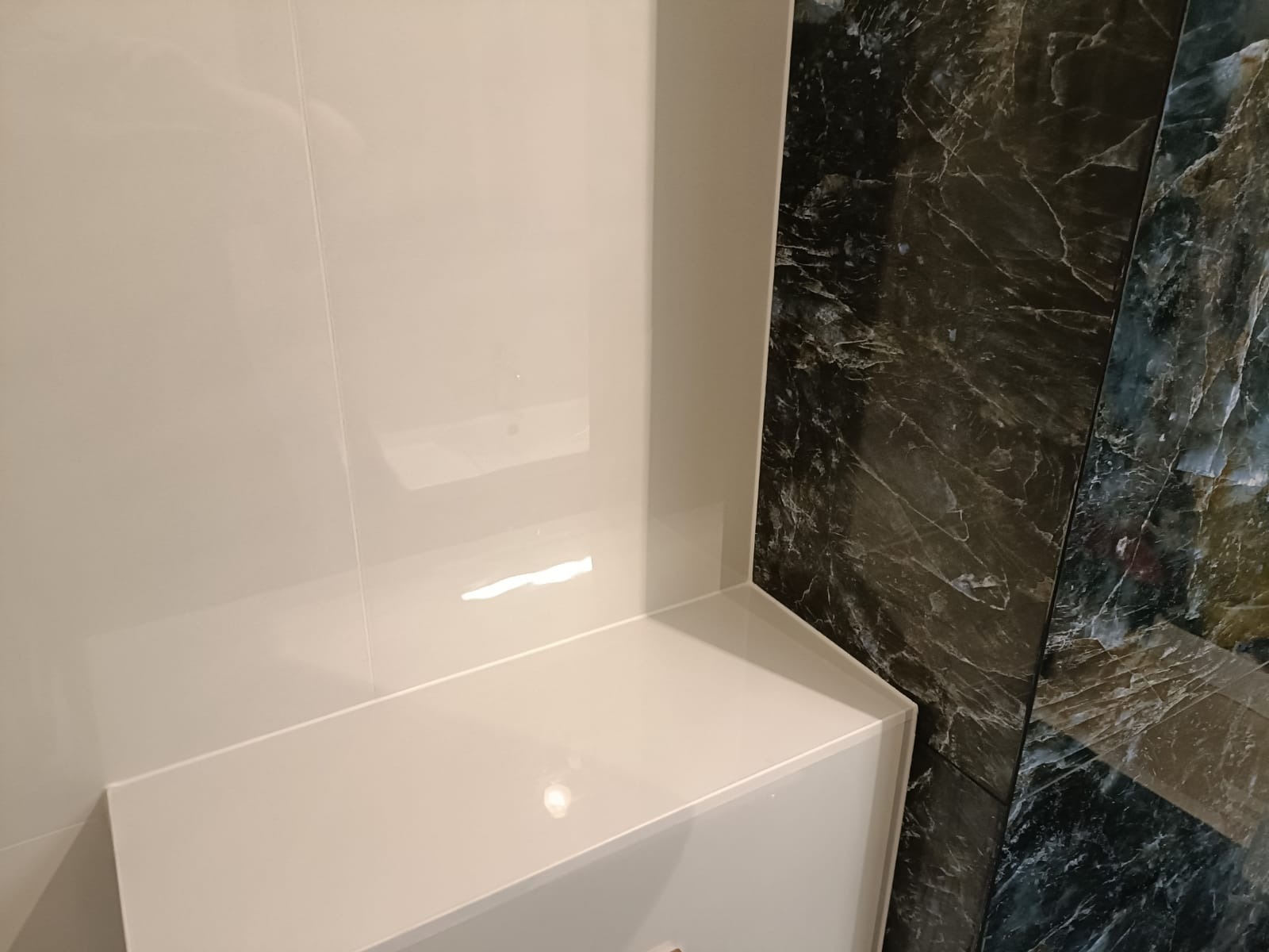 Błyszczące, białe płytki ścienne i zabudowa, kontrastują z dekoracyjną ścianą z ciemnego marmuru w łazience. Minimalistyczny design, nowoczesne wykończenie.