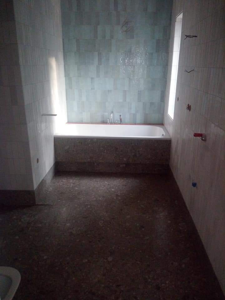 Surowa łazienka z turkusową glazurą, białą wanną i terakotą na podłodze. Widoczne wystające kable i instalacje. W oknie wpada światło dzienne.