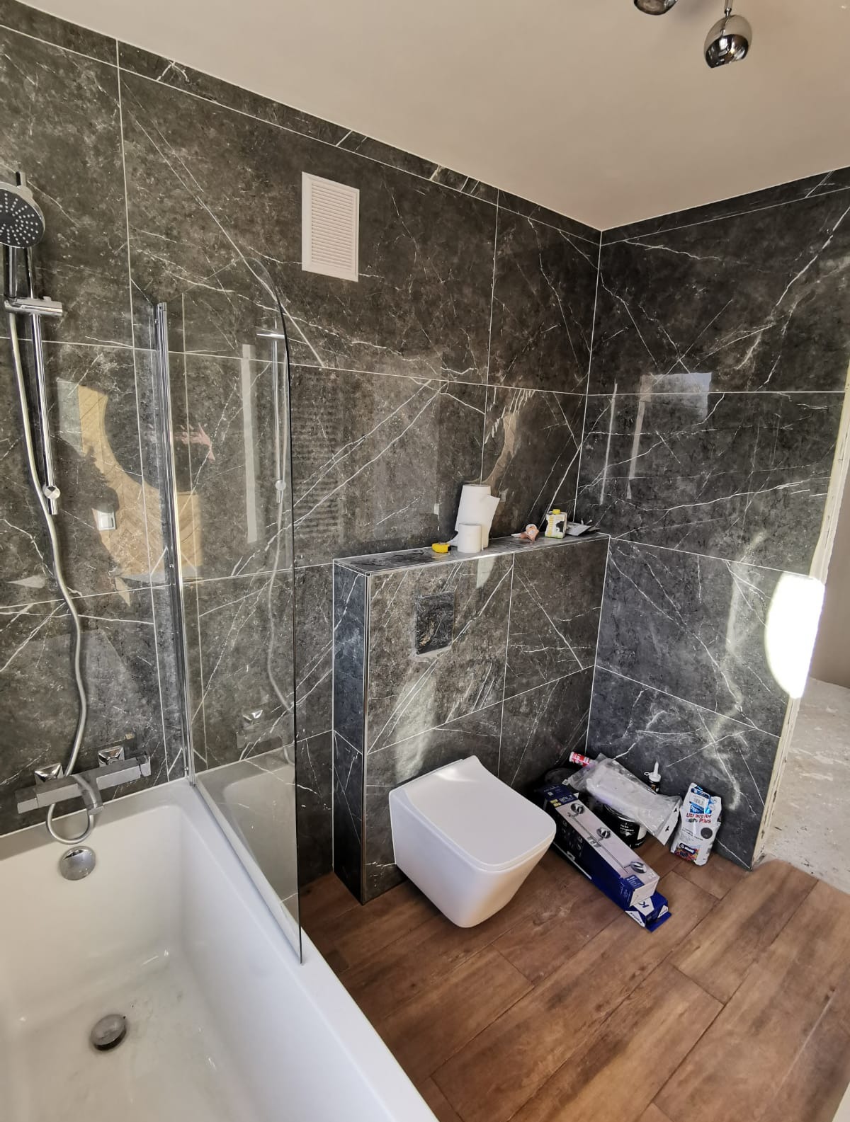 Nowoczesna łazienka w trakcie wykańczania: marmurowe płytki, biała wanna z parawanem, podwieszana toaleta i drewnopodobna podłoga. Widoczne akcesoria i narzędzia.