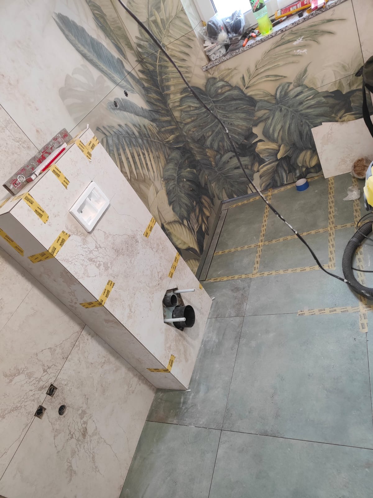 Łazienka w trakcie wykończenia: obudowa WC z płytkami, widoczne rury i odpływ liniowy, wzorzysta tapeta na ścianie z motywem roślinnym.