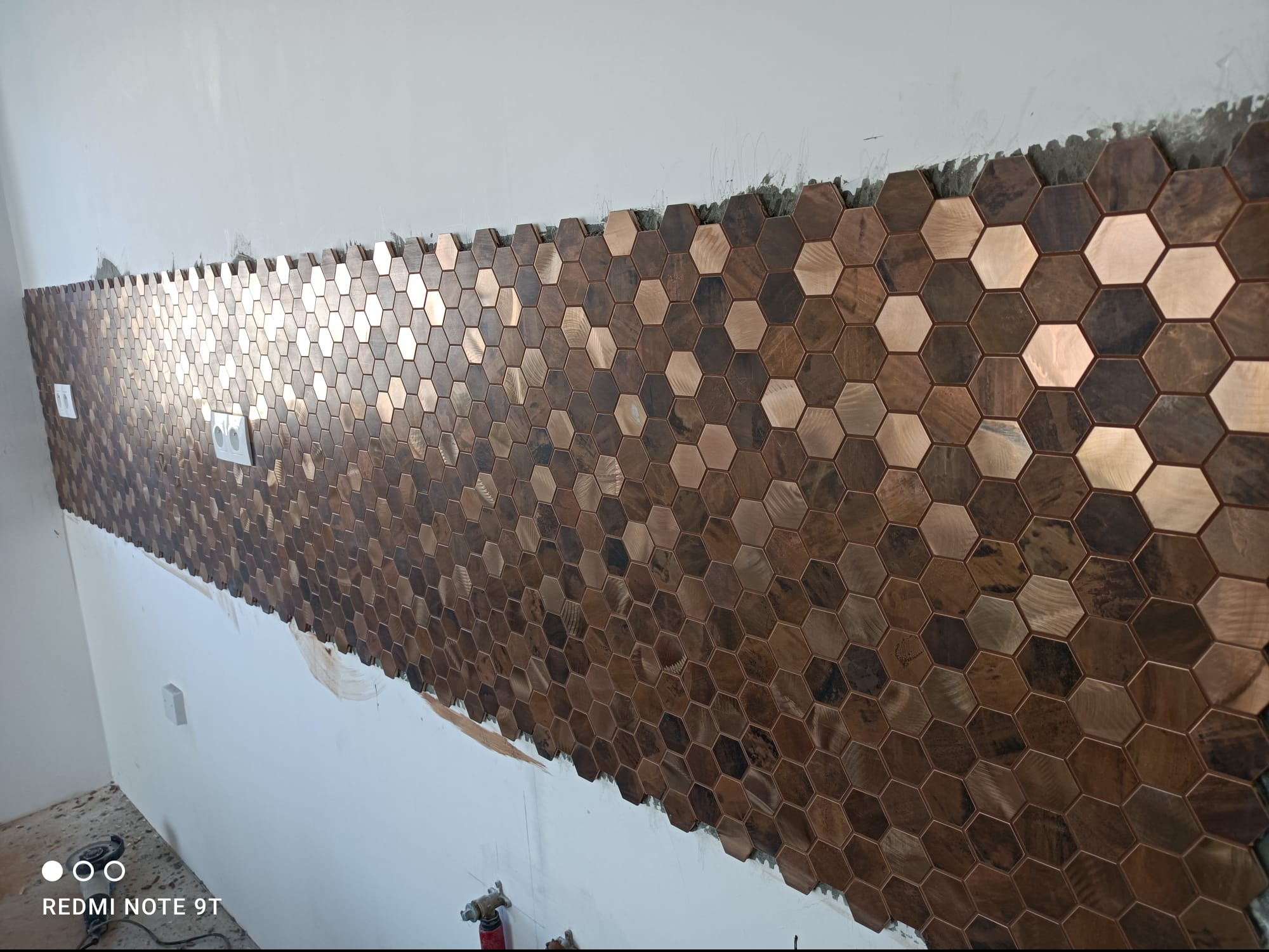 Ściana pokryta mozaiką z heksagonalnych płytek w odcieniach brązu i miedzi, z widocznymi gniazdkami elektrycznymi, częściowo wykończona, na surowym tle ściany.