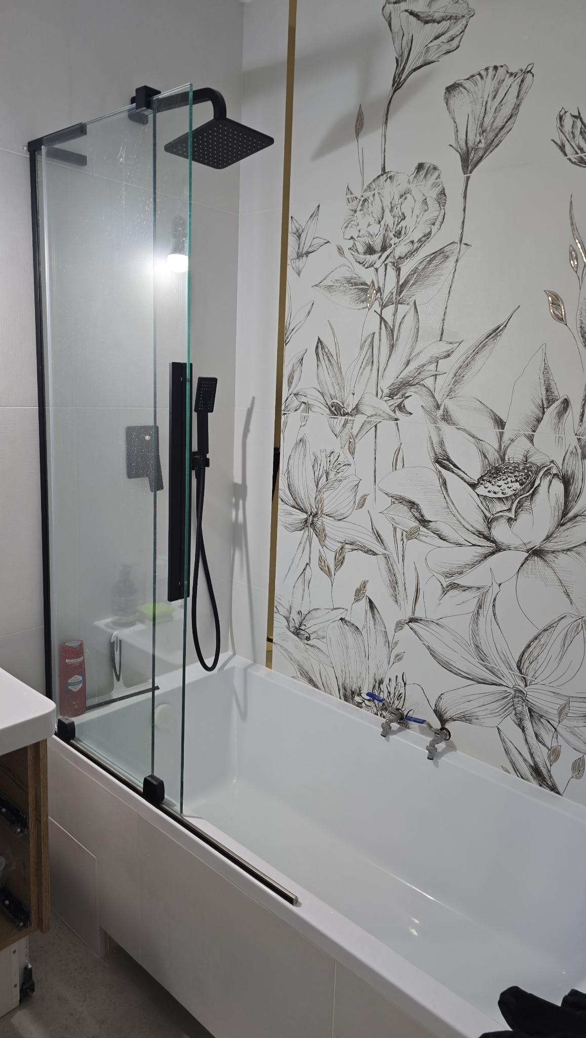 Nowoczesna łazienka z białą wanną, czarną armaturą i szklanym parawanem nawannowym. Ściana z motywem kwiatowym.