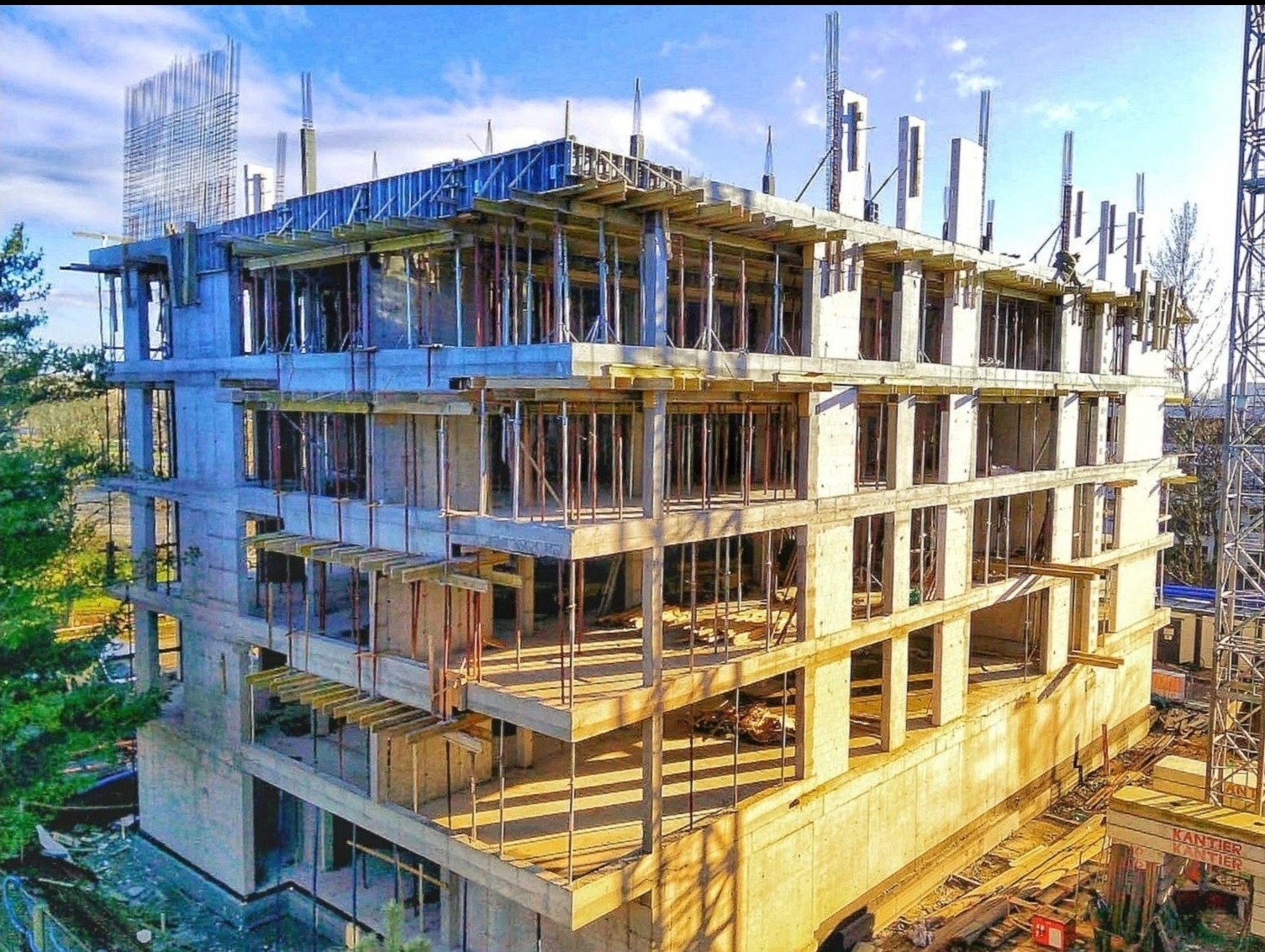 Wielopiętrowy budynek w trakcie budowy, widoczne zbrojenia, szalunki i słupy konstrukcyjne, błękitne niebo w tle. Konstrukcja w surowym stanie, bez okien i elewacji.