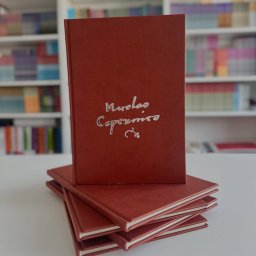 Machina Druku - Stos brązowych, oprawionych książek z tłoczonym srebrnym napisem na okładce, ustawionych na białym stole, z regałem pełnym książek w tle.