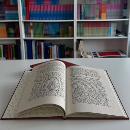 Machina Druku - Otwarta, ręcznie pisana księga w czerwonej oprawie na białym stole, z regałami pełnymi książek w tle. Widoczne ozdobne wykończenie wnętrza okładki.