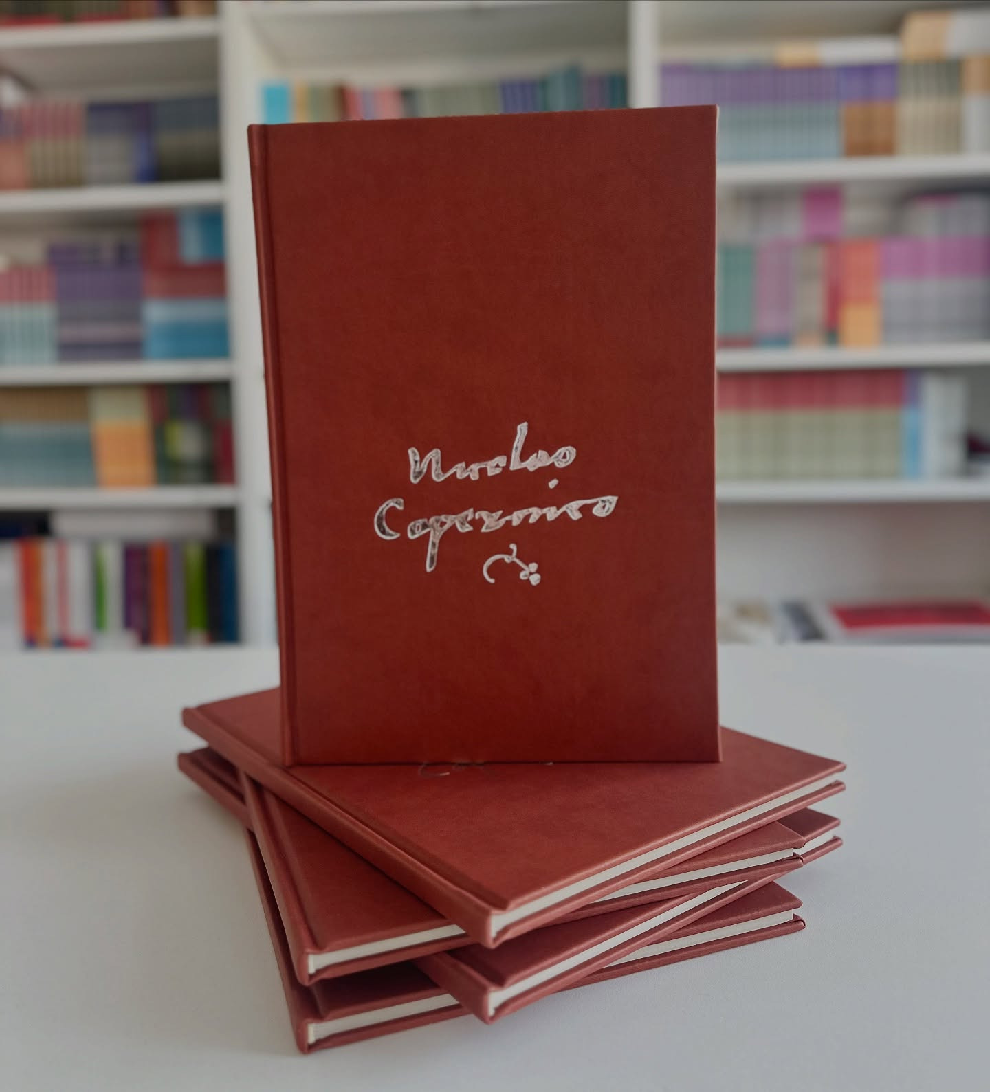 Stos brązowych, oprawionych książek z tłoczonym srebrnym napisem na okładce, ustawionych na białym stole, z regałem pełnym książek w tle.