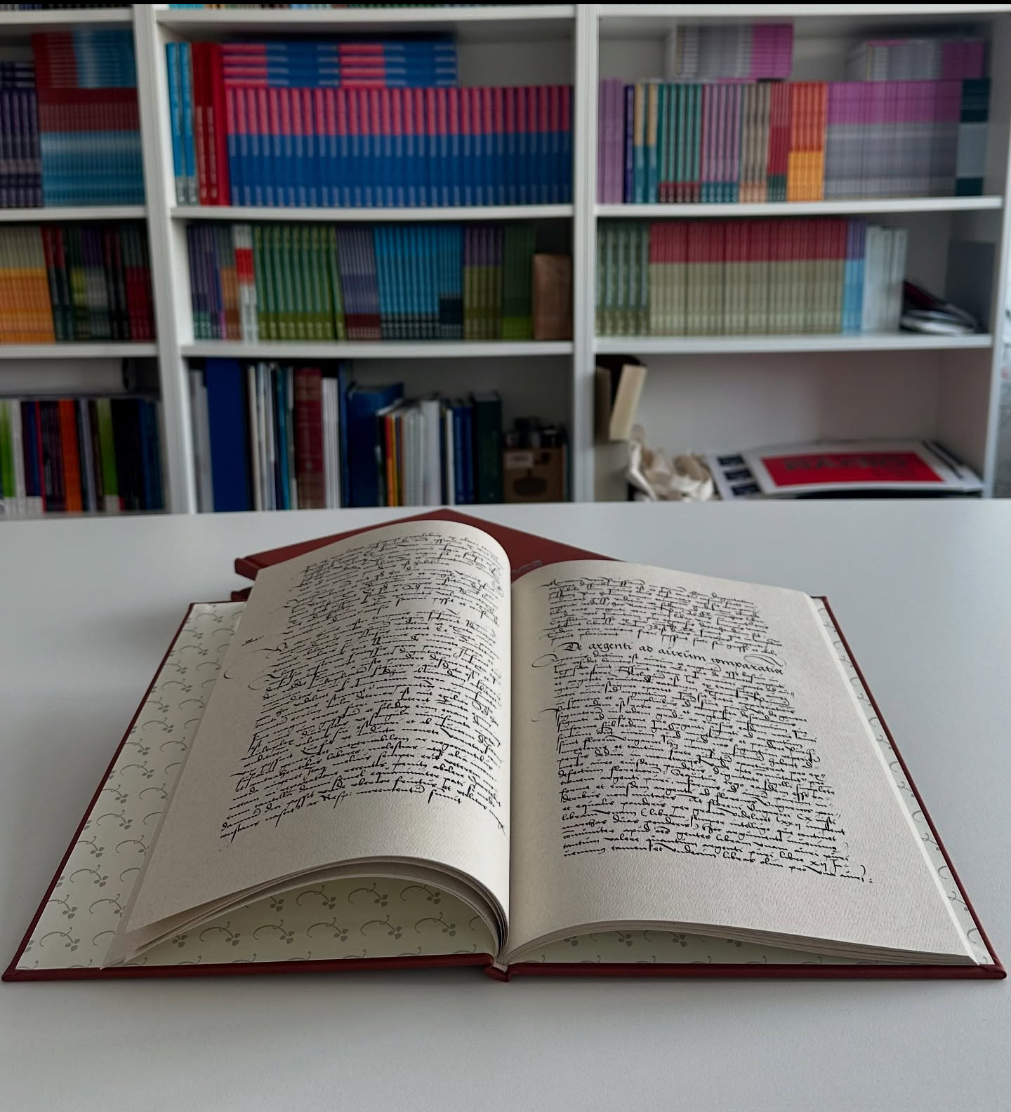 Otwarta, ręcznie pisana księga w czerwonej oprawie na białym stole, z regałami pełnymi książek w tle. Widoczne ozdobne wykończenie wnętrza okładki.