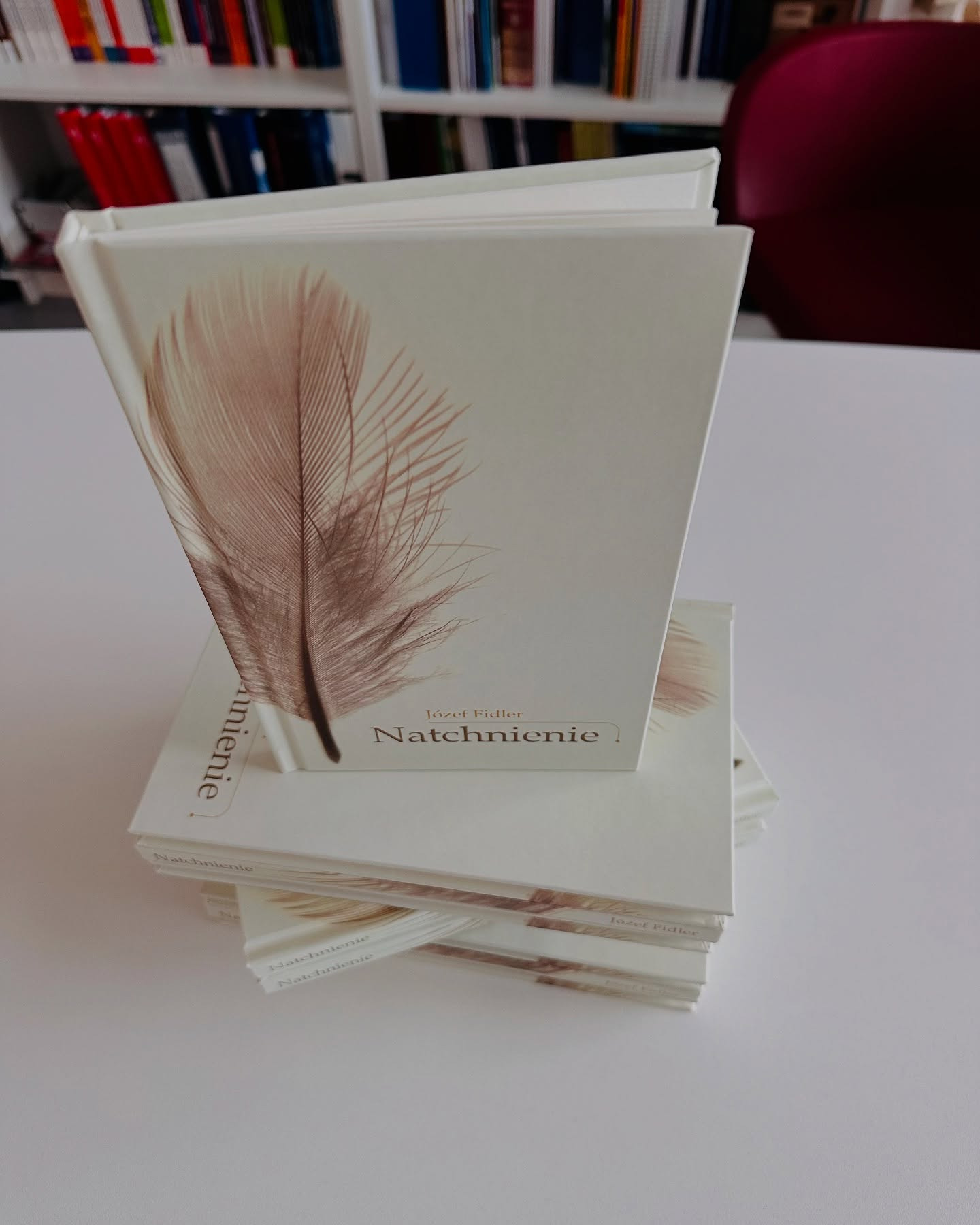 Stos książek w twardej oprawie z motywem pióra, tytułem 'Natchnienie' Józefa Fidlera, na białym stole, w tle regał z książkami i krzesło.