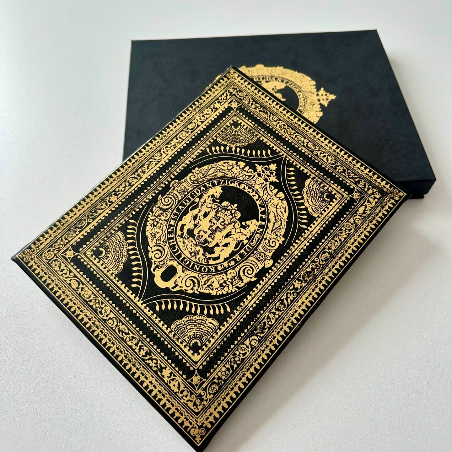 Katalog w czarnej, zdobionej złoceniami oprawie, leżący obok pasującego pudełka. Elegancki projekt graficzny z herbem i ornamentami.