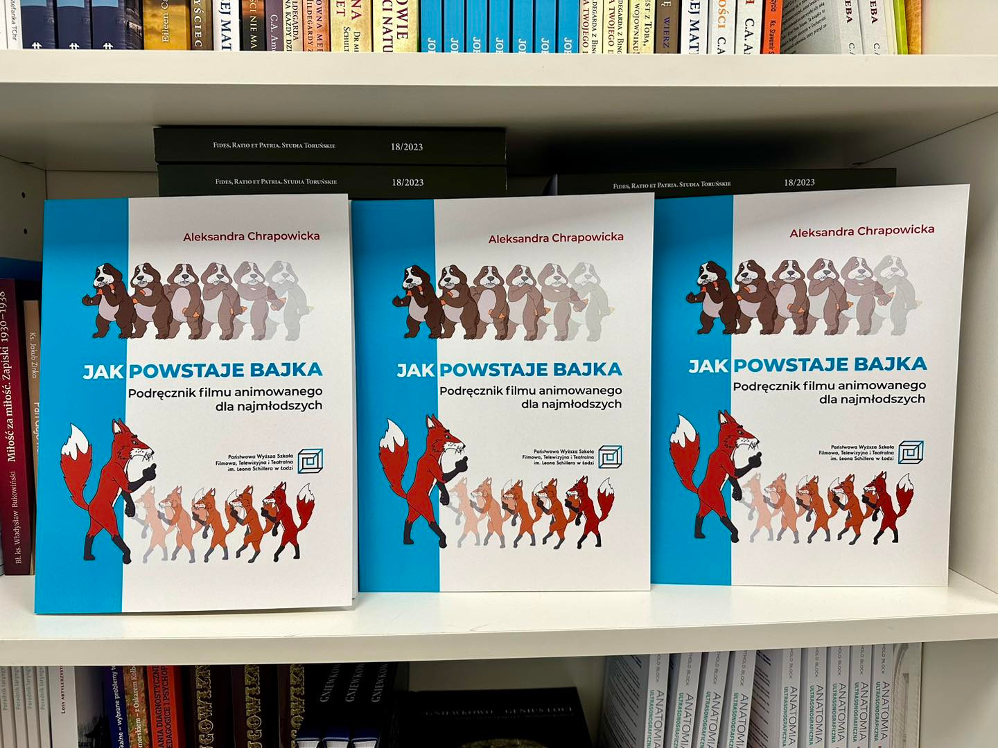 Trzy identyczne egzemplarze książki 'Jak powstaje bajka' autorstwa Aleksandry Chrapowickiej, podręcznika filmu animowanego dla najmłodszych, ustawione na półce z książkami.