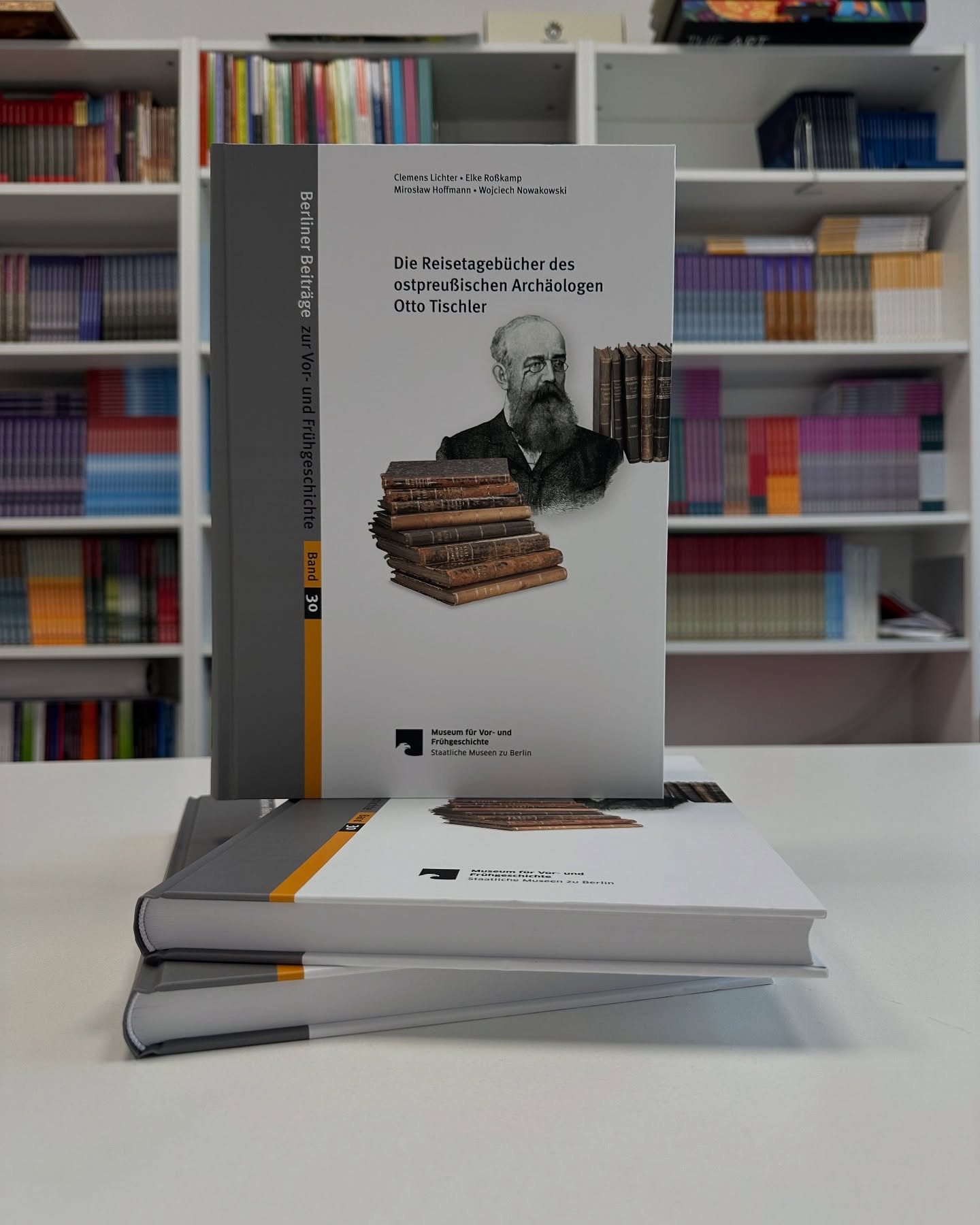 Książki z szarą okładką i żółtym paskiem na tle regału z książkami. Na okładce portret Otto Tischlera i tytuł: Die Reisetagebücher des ostpreußischen Archäologen.