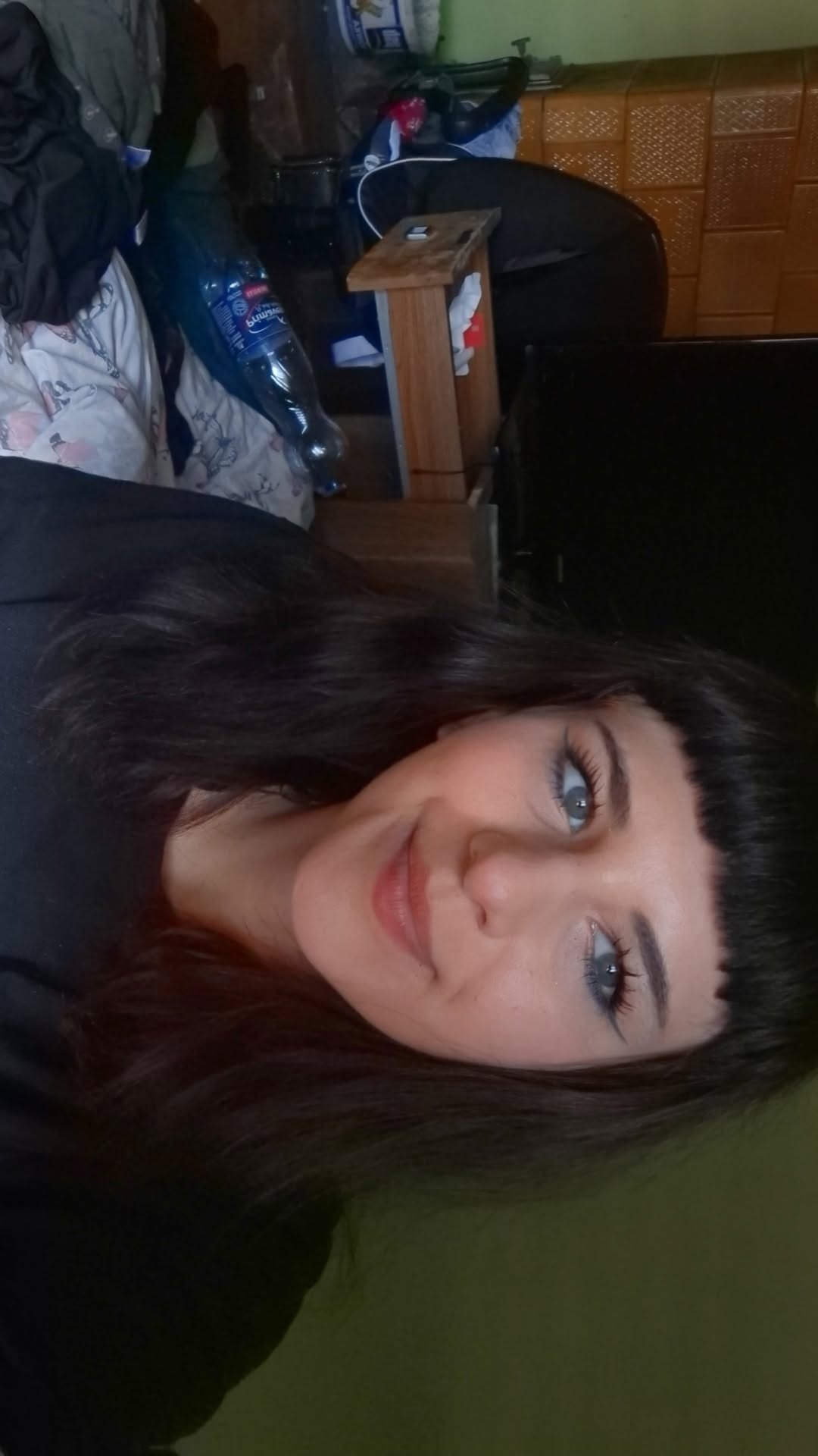 Portret uśmiechniętej kobiety z krótką grzywką na tle pokoju z widocznym łóżkiem i butelką wody. Zdjęcie z bliska.