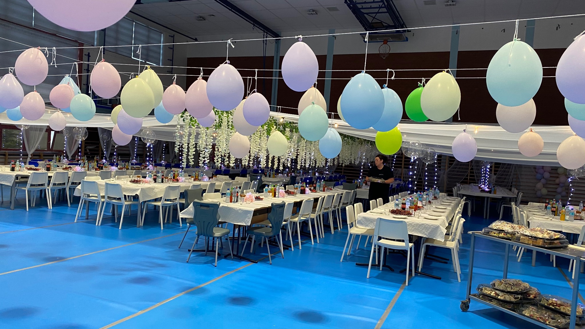 Udekorowana sala gimnastyczna na bal: pastelowe balony, białe obrusy, kwiatowe girlandy, oświetlenie punktowe, catering na wózkach. Osoba obsługuje przyjęcie.