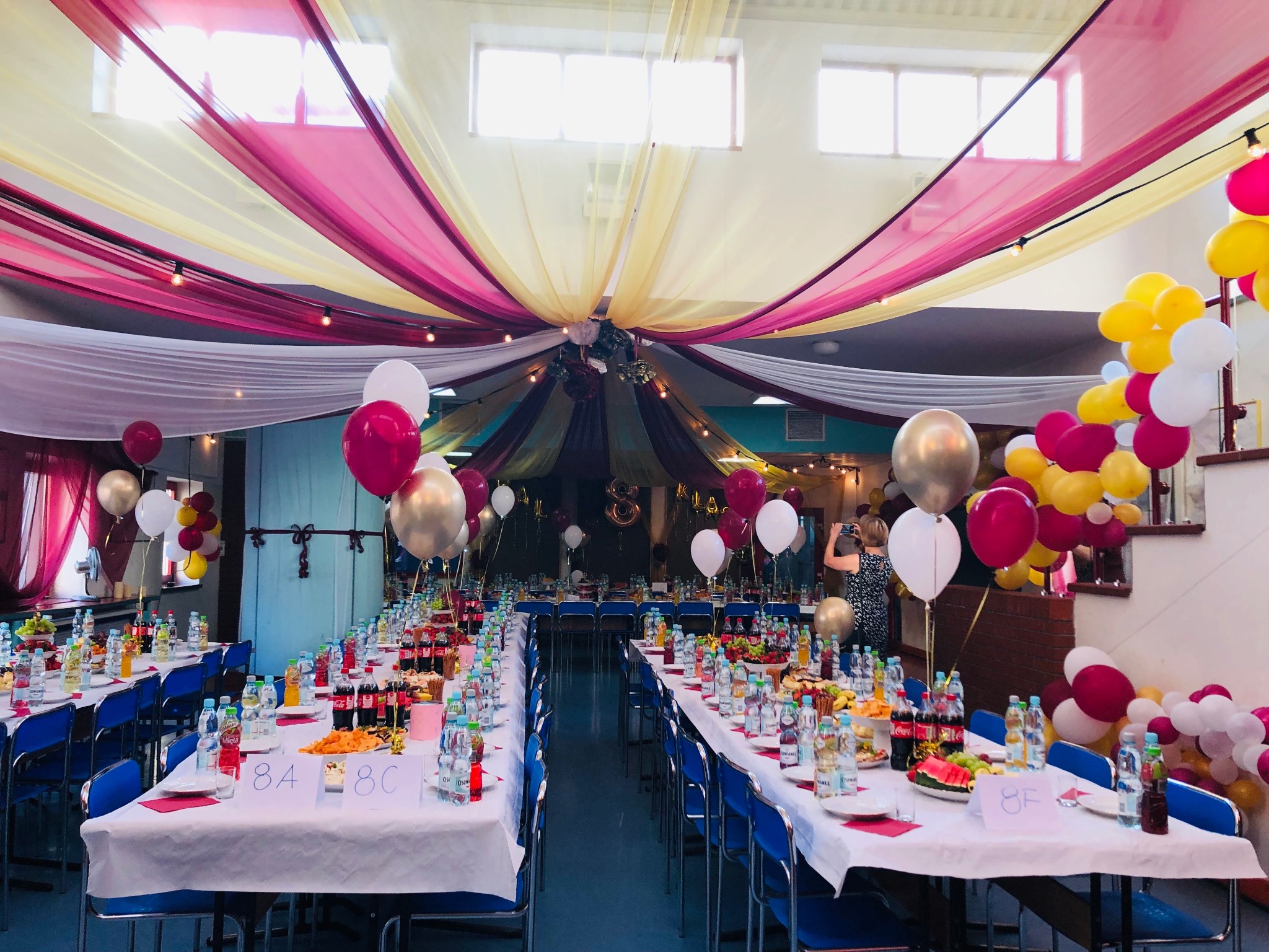 Aula szkolna udekorowana na bal 8 klasy: długie stoły z napojami i przekąskami, kolorowe balony i materiały zwisające z sufitu, atmosfera uroczystości.
