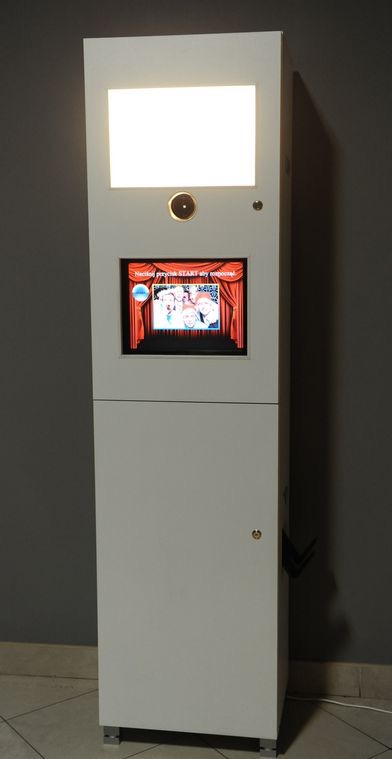 Biała fotobudka z ekranem dotykowym prezentującym zdjęcia. Górna część oświetlona, widoczne metalowe nóżki, na ekranie wyświetlona grafika z czerwonymi zasłonami.