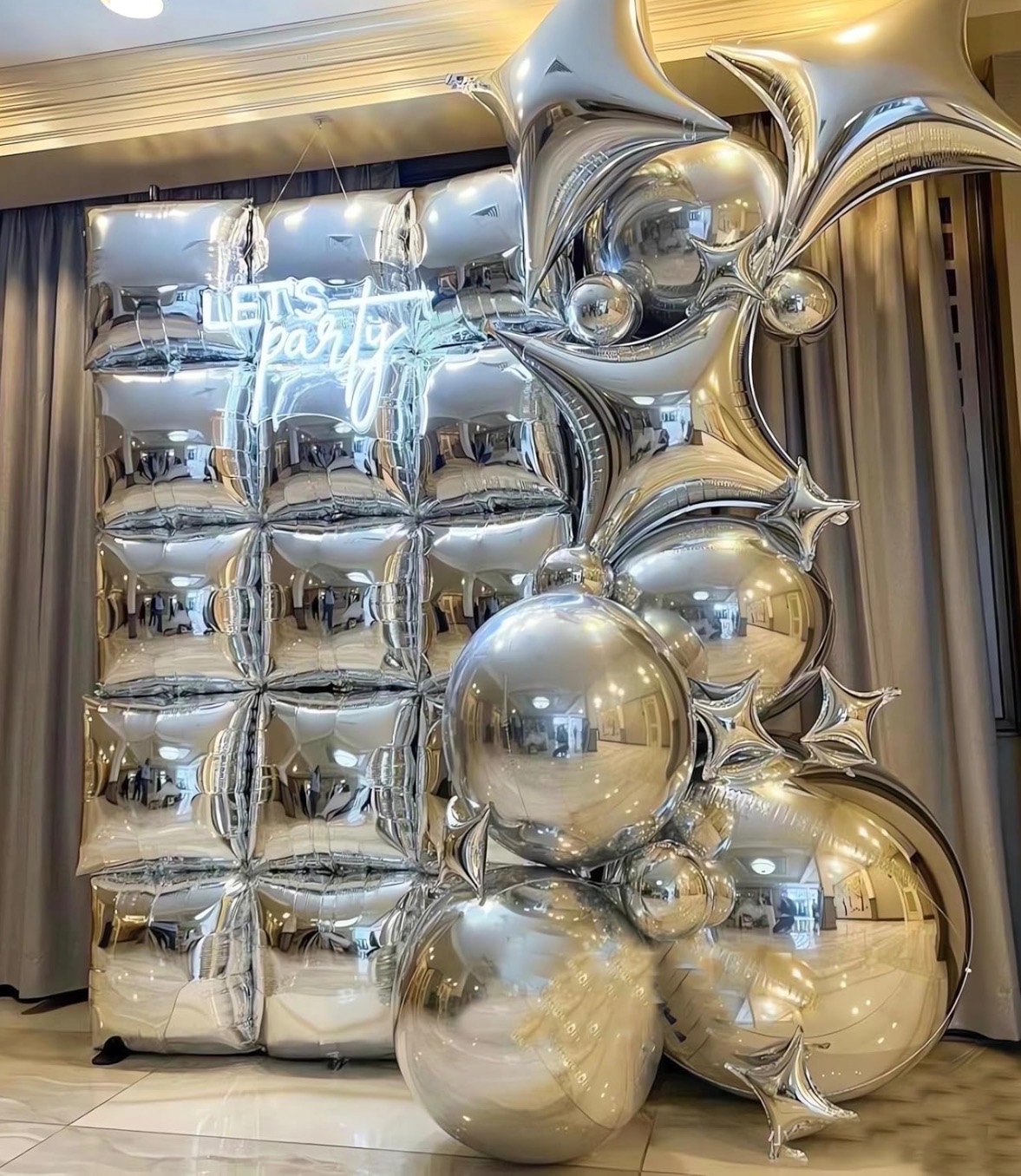 Srebrna ścianka z kwadratowych balonów i neonem 'Let's Party' oraz kompozycja z błyszczących balonów kulistych i w kształcie gwiazd, tworząca dekorację imprezową.