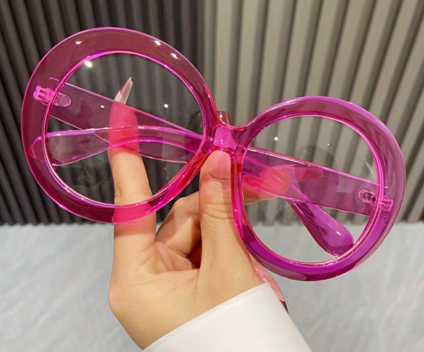 Ręka trzymająca duże, okrągłe, różowe okulary bez oprawek na rozmytym tle. Okulary mają przezroczyste szkła i plastikową ramkę w odcieniu fuksji.