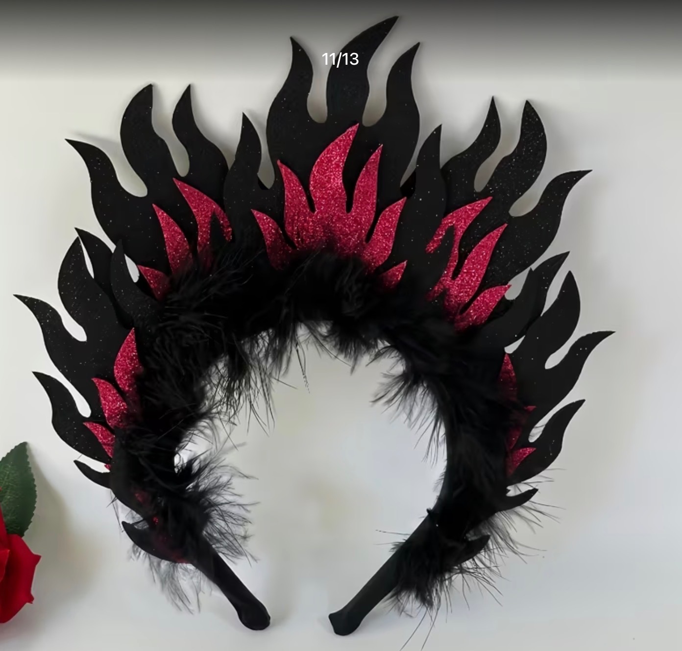 Czarno-czerwona opaska na głowę z motywem płomieni i piór, leżąca na białym tle, obok czerwonej róży. Element kostiumu scenicznego lub przebrania.