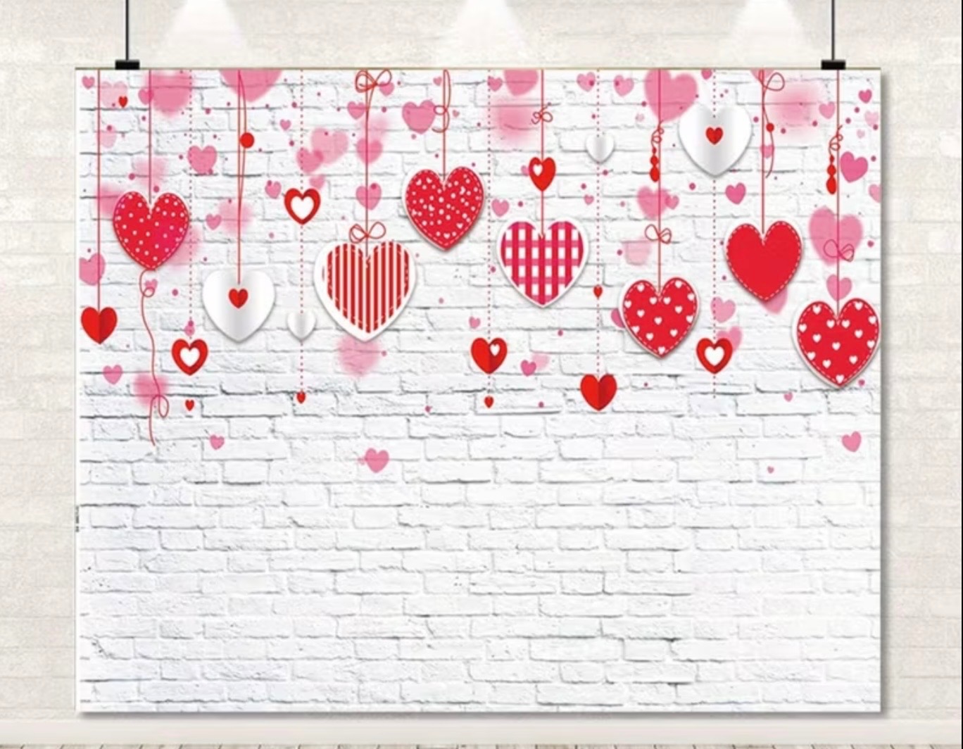 Walentynkowa ścianka fotograficzna z białej cegły, ozdobiona wiszącymi czerwonymi i białymi sercami na sznurkach, idealna na sesje zdjęciowe.