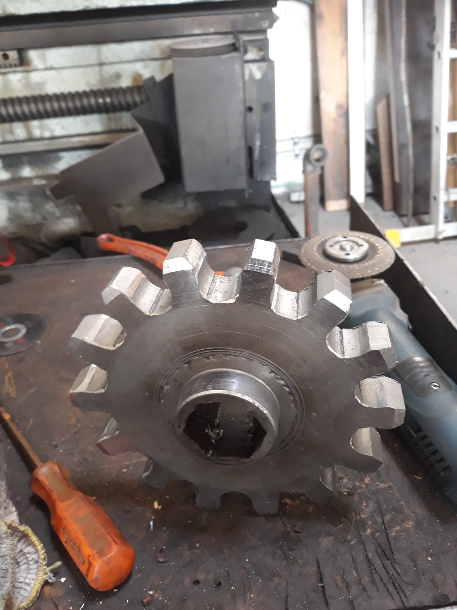 Metalowa zębatka z sześciokątnym otworem, leżąca na warsztatowym stole z narzędziami. Widoczne ślady obróbki.