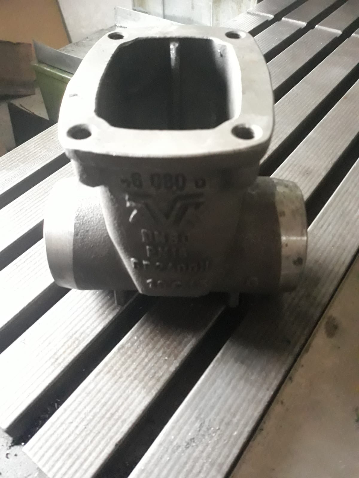 Szara, metalowa kształtka z czterema otworami na kołnierzu, leżąca na metalowym stole z rowkami. Widoczne napisy i oznaczenia na korpusie.