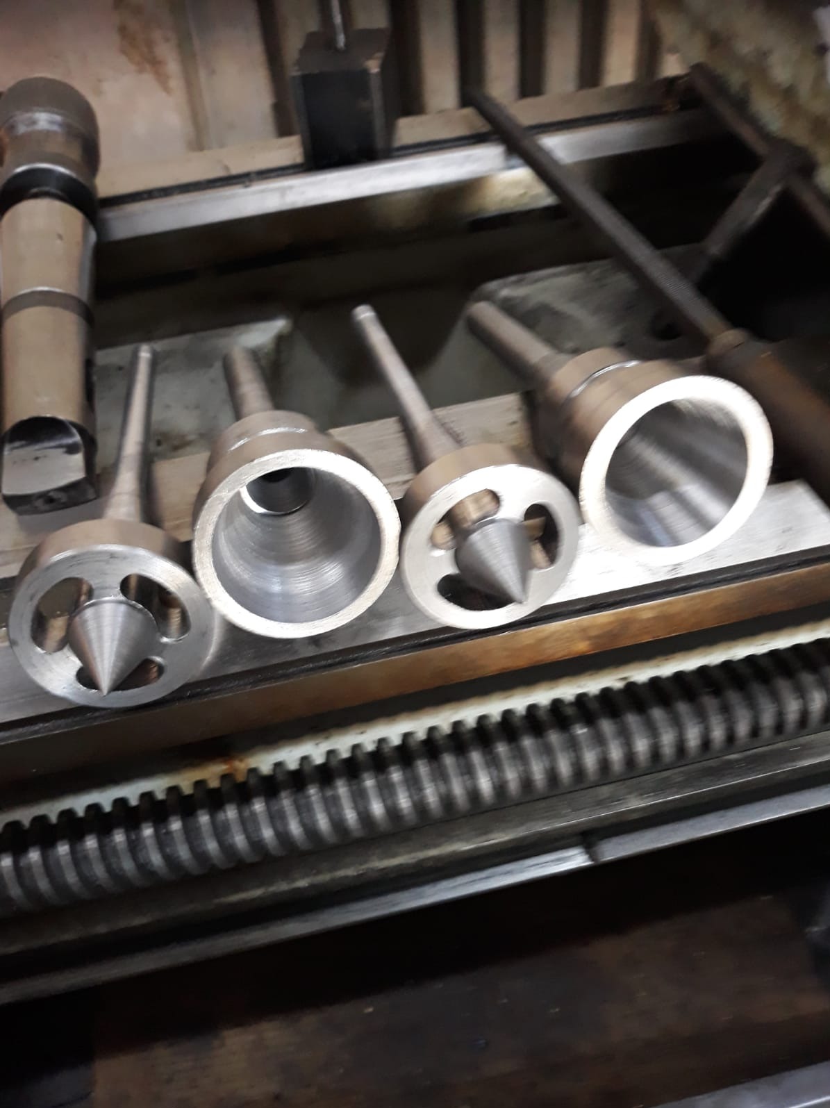 Metalowe narzędzia tokarskie o różnych kształtach i rozmiarach ułożone na metalowej powierzchni tokarki. Widoczne elementy tokarki w tle.