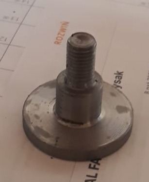 Metalowy element z gwintem i okrągłą podstawą, po obróbce tokarskiej, widoczne ślady spawania, na tle kartki z notatkami, lekko zamazane krawędzie.