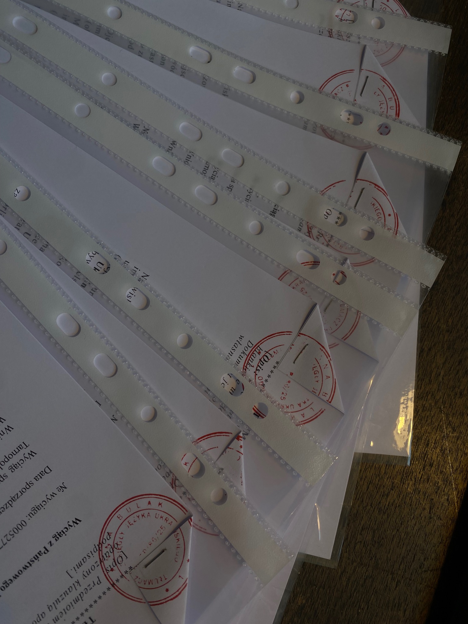 Stos dokumentów z pieczątkami tłumacza przysięgłego w foliowych koszulkach, ułożonych pod kątem na drewnianym podłożu. Widoczne fragmenty treści w języku ukraińskim.