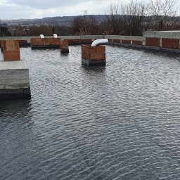 POL-KRAJ DACHY PŁASKIE PIOTR KRAJEWSKI - Płaski dach pokryty wodą po deszczu, z widocznymi kominami i obróbkami blacharskimi. W tle zabudowa miejska i wzgórza.