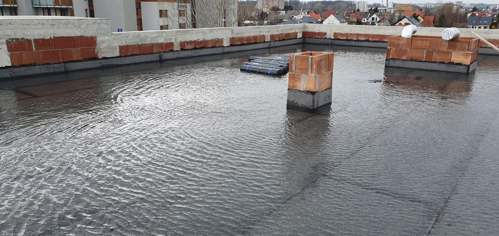 Zalany, płaski dach po wykonaniu hydroizolacji z papy. Widoczny komin z cegły i rolki papy. W tle zabudowa miejska.