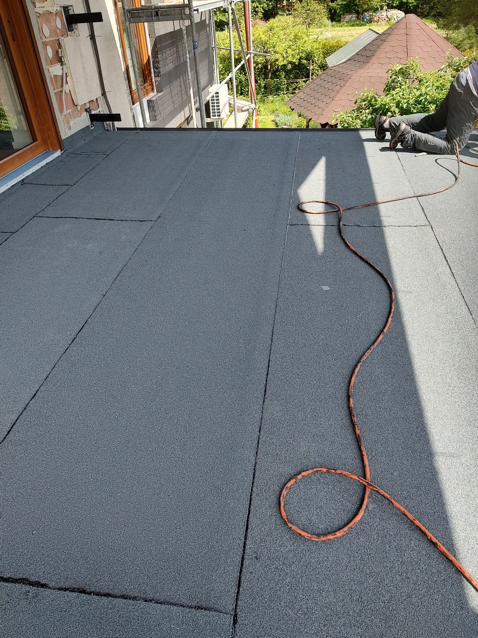Szary dach pokryty papą termozgrzewalną w trakcie prac dekarskich; widoczny fragment dachu i pracujący dekarz. Na dachu leży pomarańczowy kabel zasilający.